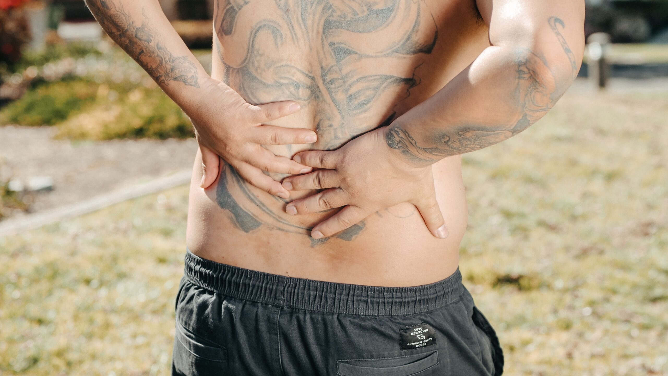 A man with a large tattoo on his back holds his lower back outdoors, showing discomfort.